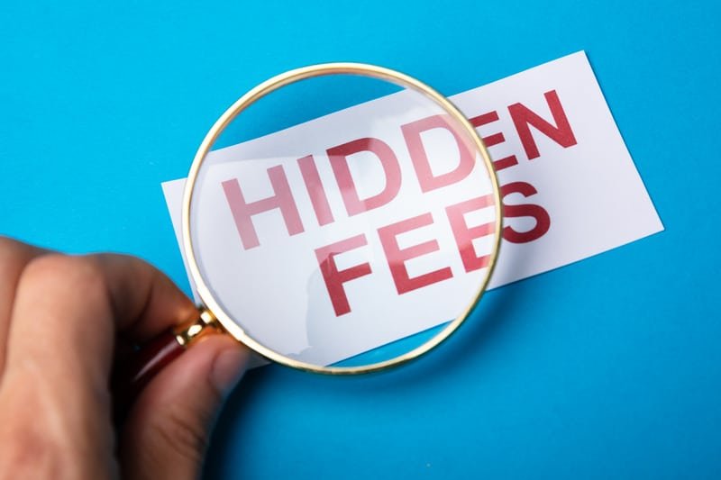 the words Hidden Fees appear on a paper. The letters are capitalized and in red. A hand holding a magnifying glass hovers over the words.